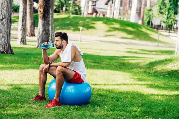 Barbudo deportista beber agua de deporte botella mientras está sentado en la pelota de fitness - foto de stock