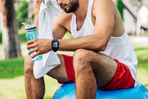 Vista recortada del hombre barbudo sosteniendo la botella deportiva mientras está sentado en la pelota de fitness y limpiando el sudor - foto de stock
