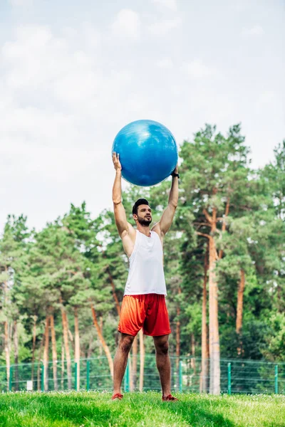 Apuesto deportista haciendo ejercicio mientras sostiene la pelota de fitness en el parque - foto de stock