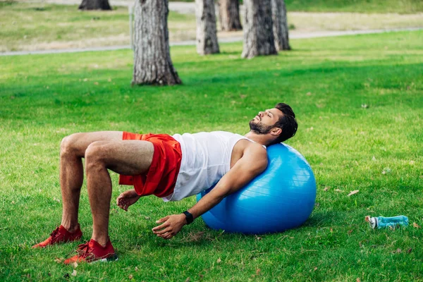 Bell'uomo barbuto che lavora sulla palla fitness vicino alla bottiglia di sport — Foto stock