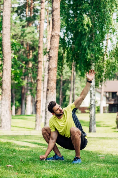 Красивий молодий спортсмен сидить на траві і махає рукою в парку — стокове фото