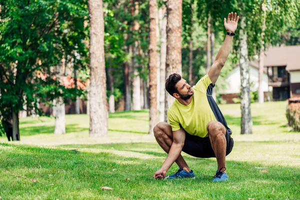 Guapo joven deportista sentado en la hierba y agitando la mano - foto de stock