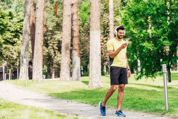 Felice uomo barbuto utilizzando smartphone e ascoltare musica in cuffia mentre si cammina nel parco — Foto stock