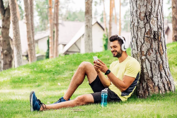 Homem alegre em fones de ouvido usando smartphone perto árvore tronco e ouvir música — Fotografia de Stock