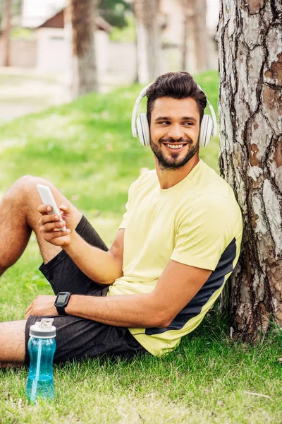 Homem positivo em fones de ouvido usando smartphone perto árvore tronco e ouvir música — Fotografia de Stock