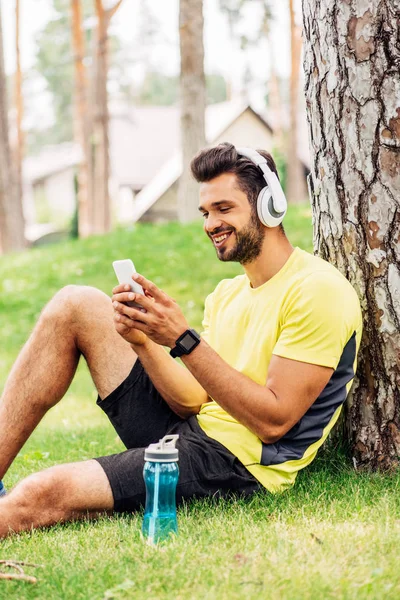 Happy sportsman in headphones using smartphone near tree trunk and listening music — Stock Photo