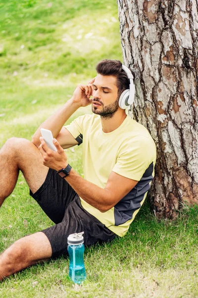 Schöner Sportler mit Kopfhörer, der sein Smartphone in der Nähe des Baumstammes benutzt und Musik hört — Stockfoto