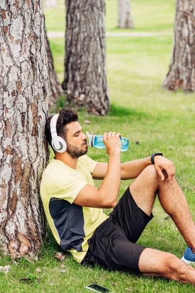 Belo desportista em fones de ouvido beber água perto de árvore tronco — Fotografia de Stock