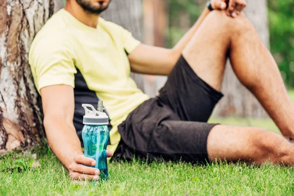 Vue recadrée d'un sportif barbu tenant une bouteille de sport près d'un tronc d'arbre — Photo de stock