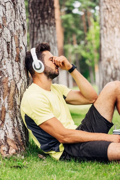 Sportsman in headphones covering face while holding sport bottle near tree trunk — Stock Photo