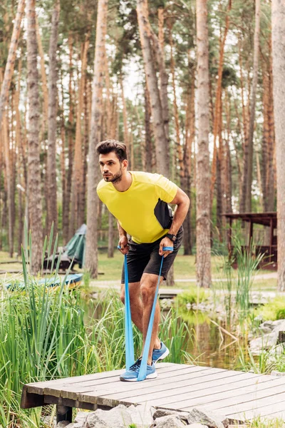 Homem bonito exercitando com tiras de suspensão perto do lago na floresta — Fotografia de Stock