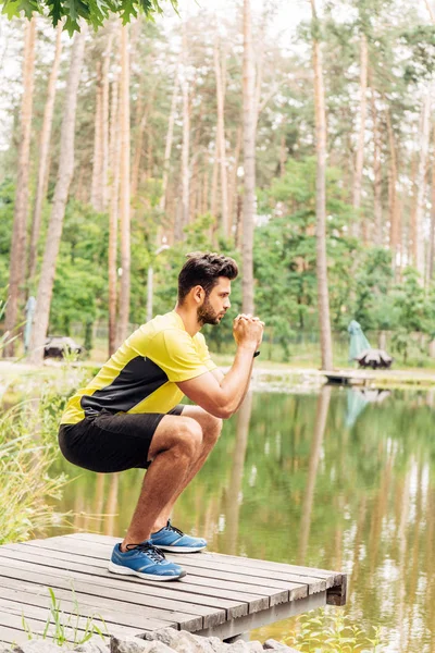 Athletic sportsman in sportswear doing exercise near lake in forest — Stock Photo