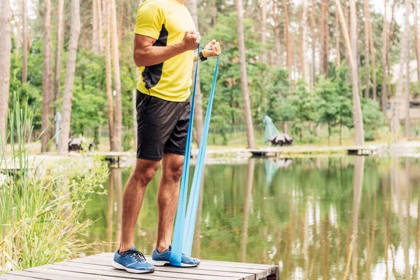 Vista recortada de deportista haciendo ejercicio con correas de suspensión cerca del lago - foto de stock