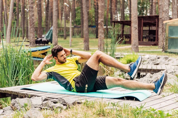 Apuesto deportista de entrenamiento en la alfombra de fitness cerca de piedras y árboles - foto de stock