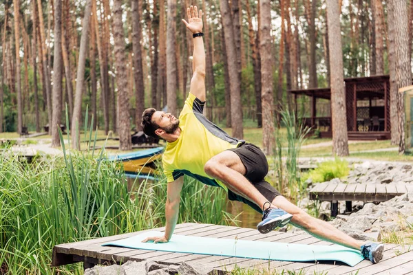 Entrenamiento de deportista barbudo en colchoneta de fitness cerca de piedras y árboles - foto de stock