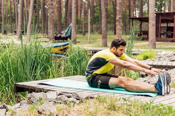 Bearded man in sportswear stretching on fitness mat near stones and trees — Stock Photo