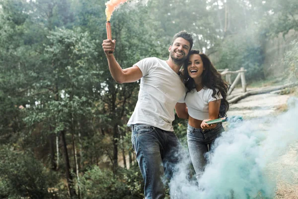 Atractiva mujer y hombre barbudo alegre sosteniendo bombas de humo - foto de stock