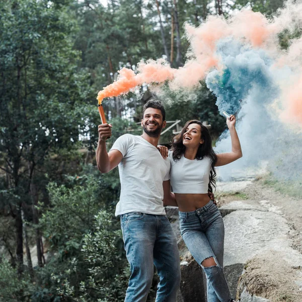 Atractiva mujer y hombre barbudo alegre sosteniendo bombas de humo fuera - foto de stock
