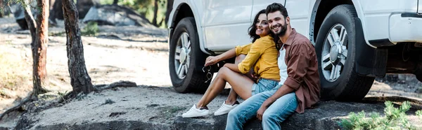 Tiro panorámico de hombre barbudo feliz y chica atractiva sentada cerca de coche y árboles - foto de stock