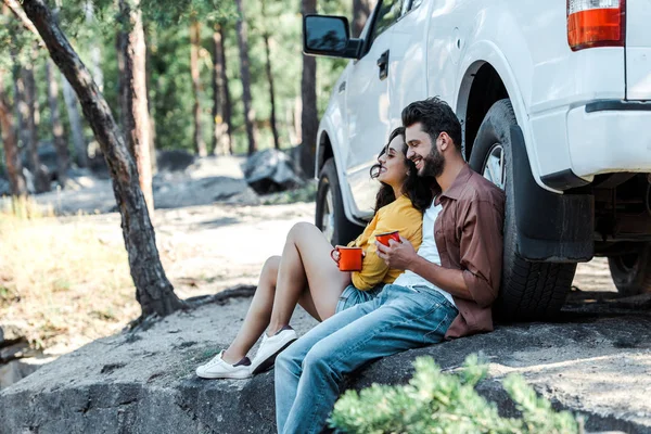 Enfoque selectivo de hombre y mujer feliz sosteniendo tazas mientras está sentado cerca de coche en el bosque - foto de stock