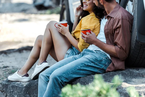 Abgeschnittene Ansicht eines bärtigen Mannes und eines glücklichen Mädchens, die neben dem Auto sitzen und Tassen in der Hand halten — Stockfoto