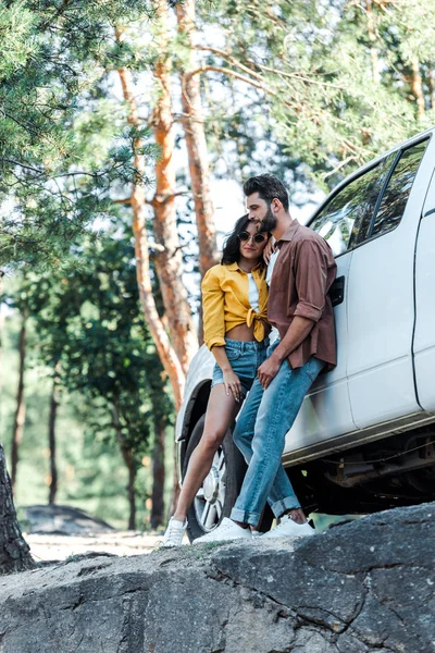 Felice uomo barbuto e allegra ragazza in occhiali da sole in piedi vicino auto nel bosco — Foto stock