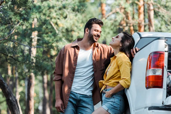 Hombre barbudo feliz de pie y mirando a la chica sonriendo cerca del coche - foto de stock