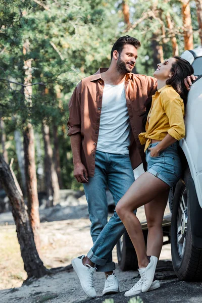 Hombre barbudo guapo de pie y mirando a la chica sonriendo cerca del coche - foto de stock