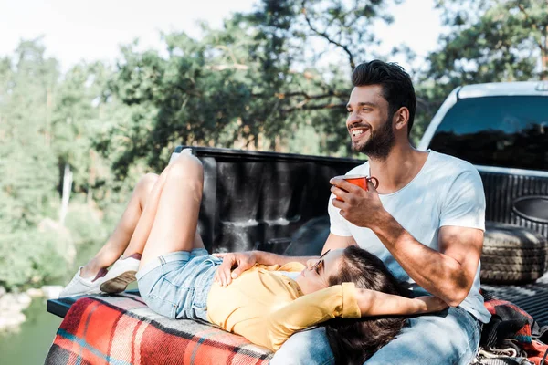 Handsome bearded man smiling and holding cup while girl lying on plaid blanket — Stock Photo