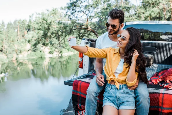 Веселая девушка в солнечных очках говорит селфи с мужчиной возле машины и озера — стоковое фото