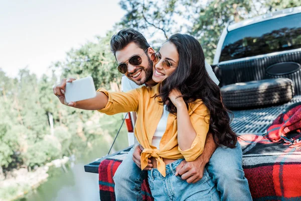 Alegre chica en gafas de sol hablando selfie con barbudo hombre cerca de coche - foto de stock