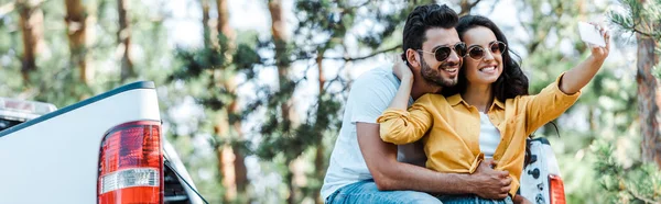 Plan panoramique de fille gaie parler selfie avec l'homme près de la voiture et les arbres — Photo de stock