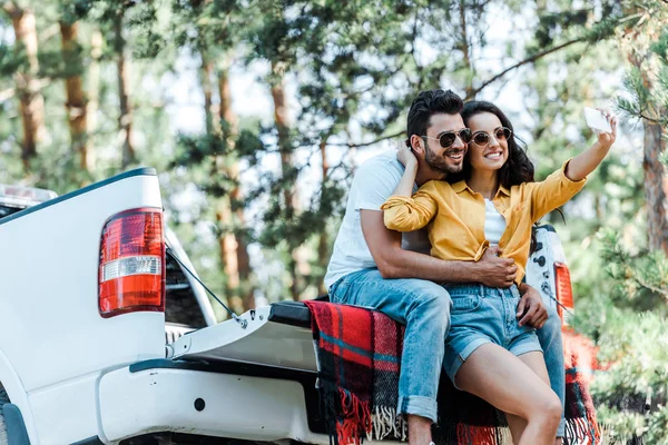 Cheerful young woman talking selfie with bearded man near car and trees — Stock Photo