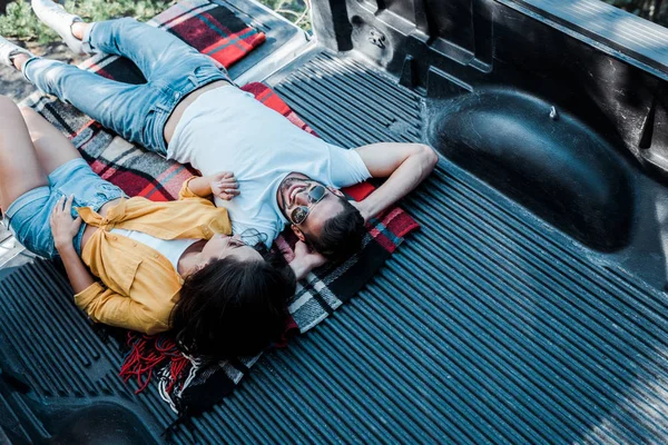 Overhead view of woman in sunglasses lying with happy man in plaid blanket in car trunk — Stock Photo