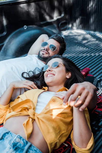 Woman in sunglasses lying with happy man in plaid blanket in car trunk — Stock Photo