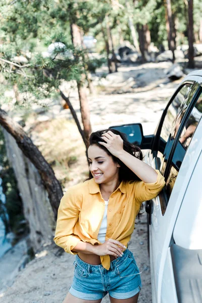 Foco seletivo da mulher atraente sorrindo enquanto está perto do carro em madeiras — Fotografia de Stock