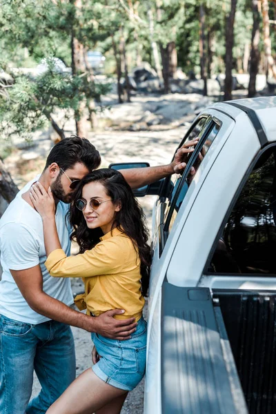 Menina atraente em óculos de sol abraçando homem barbudo perto do carro — Fotografia de Stock