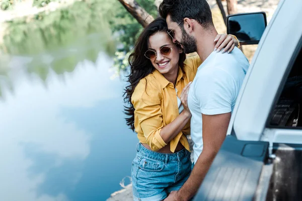 Enfoque selectivo de la chica feliz abrazo con novio cerca de coche y río - foto de stock