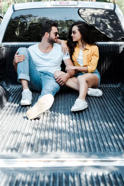 Cheerful man in sunglasses sitting in car trunk and looking at girl — Stock Photo