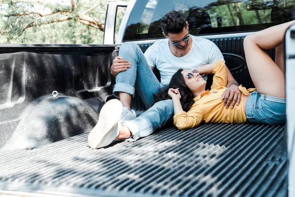 Hombre barbudo en gafas de sol sentado en el maletero del coche y mirando a la chica - foto de stock