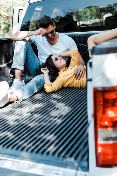 Вибірковий фокус бородатого чоловіка в сонцезахисних окулярах, сидячи в багажнику автомобіля і дивлячись на дівчину — стокове фото