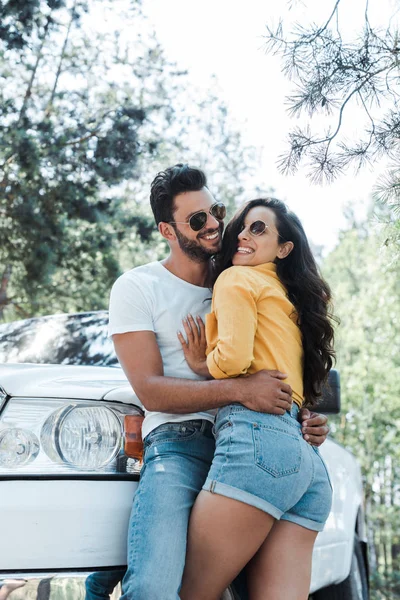 Vista basso angolo di uomo in piedi e abbracciando donna felice vicino auto nei boschi — Foto stock