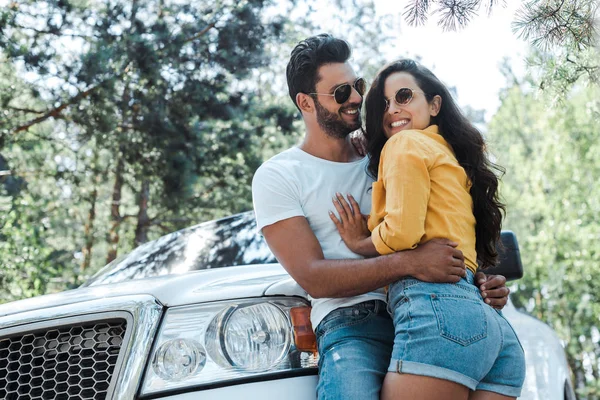 Visão de baixo ângulo do homem de pé e abraçando menina feliz perto de automóvel na floresta — Fotografia de Stock