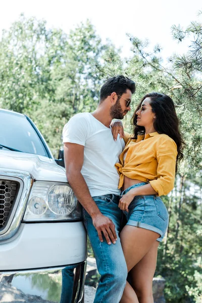 Hombre guapo de pie y abrazando chica feliz cerca de auto en el bosque - foto de stock