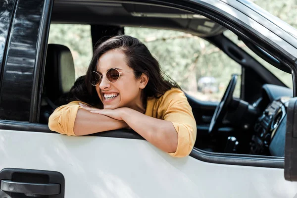 Chica alegre en gafas de sol sonriendo desde la ventana del coche - foto de stock