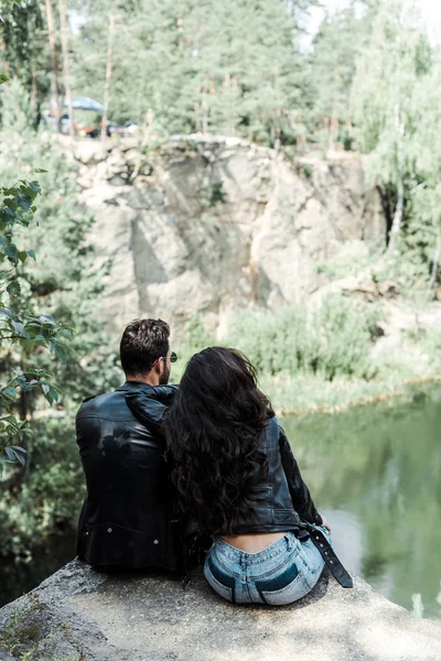 Visão traseira de homem e mulher em jaquetas de couro sentado perto de árvores em bosques — Fotografia de Stock