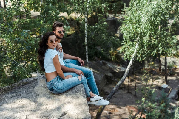 Hombre guapo y mujer atractiva en gafas de sol sentado cerca de los árboles — Stock Photo