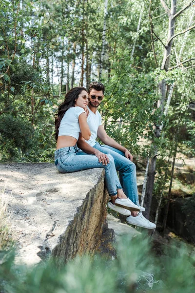 Messa a fuoco selettiva di bell'uomo e donna attraente in occhiali da sole seduti vicino agli alberi con foglie — Foto stock