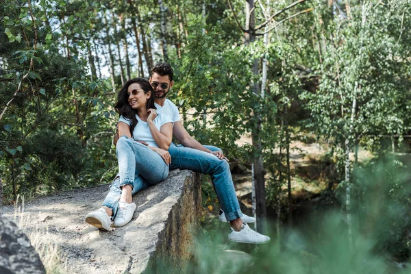 Selektiver Fokus eines gutaussehenden Mannes und einer fröhlichen Frau mit Sonnenbrille, die neben Bäumen mit Blättern sitzen — Stockfoto