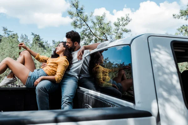 Vue à angle bas de bel homme barbu assis sur la voiture et regardant jeune femme heureuse — Photo de stock